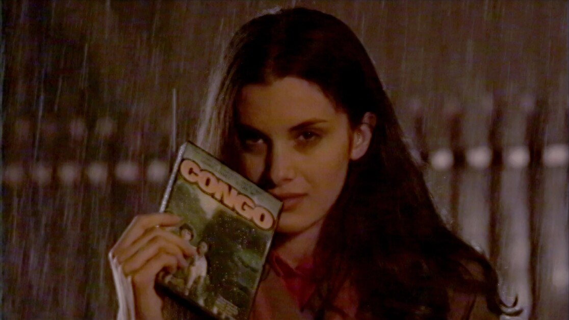 A person with long dark hair stands in the rain at night, holding a copy of the movie "Congo" up to their face.