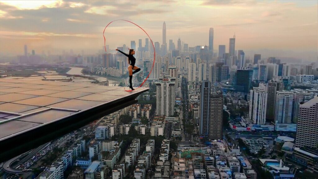 A person performs a high-leg kick on the edge of a rooftop with a red ribbon, against the backdrop of a sprawling cityscape at sunset.