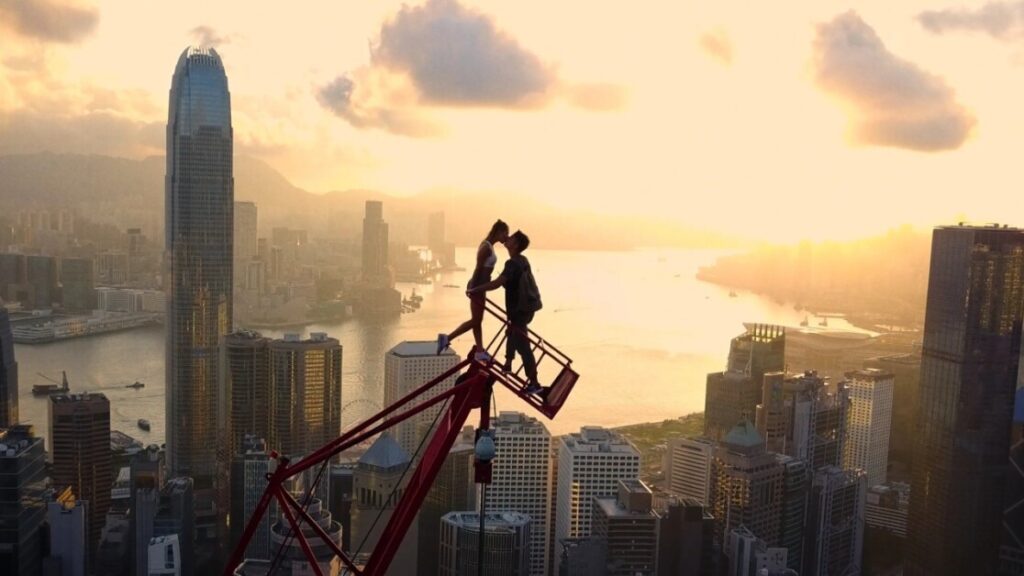 Two people are kissing while standing on top of a tall antenna with a city skyline and a large body of water in the background during sunset.