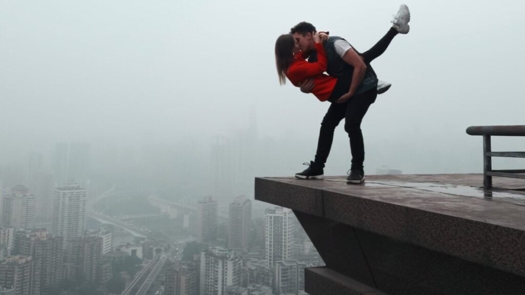 A couple stands on the edge of a tall building, with the woman lifted in the air as they kiss, overlooking a foggy cityscape.