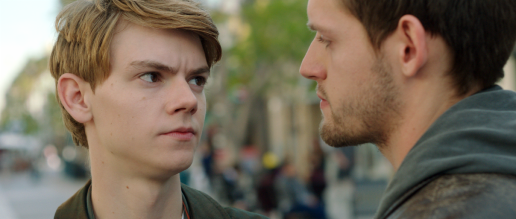 Two young men stand facing each other outdoors; one looks serious while the other appears calm.