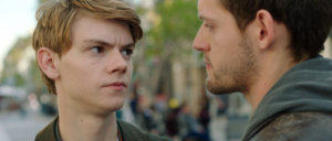 Two young men stand facing each other outdoors; one looks serious while the other appears calm.