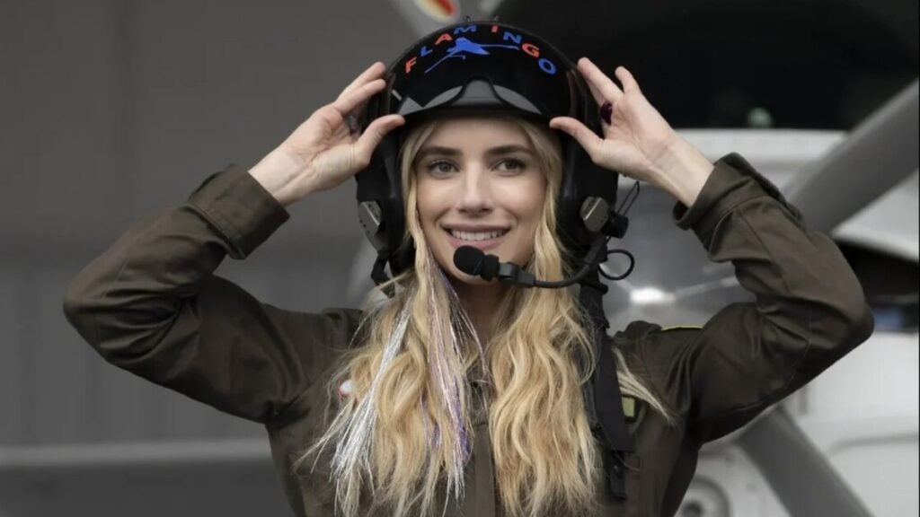 A person wearing a dark flight suit and helmet smiles while standing in front of an aircraft. They have long blonde hair and a communication headset attached to the helmet.