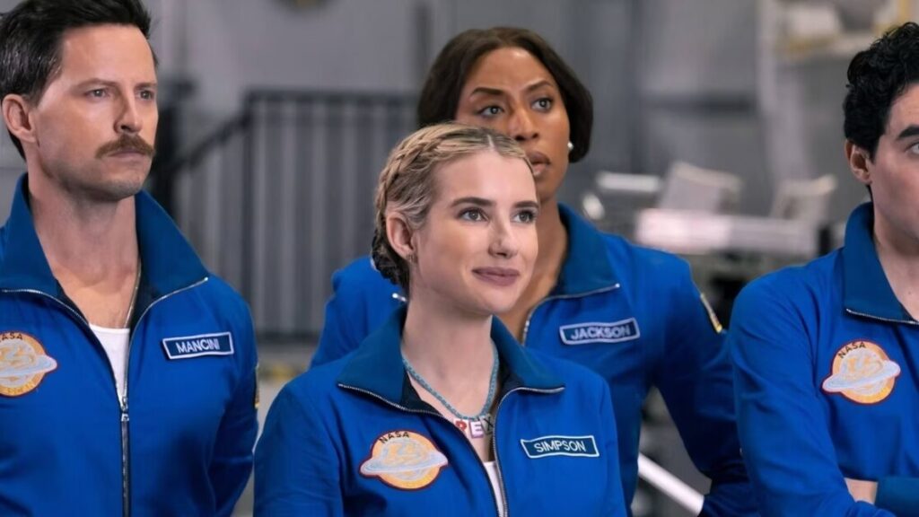 Four individuals wearing blue space suits stand together, each with a name patch (Mancon, Simpson, Jackson). The background is an indoor setting with blurred equipment.