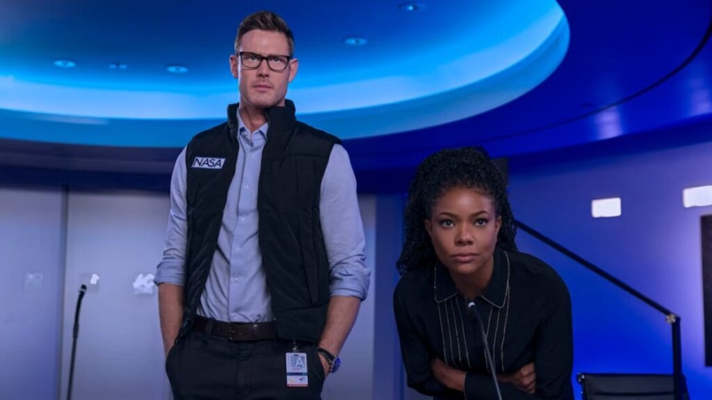 Two individuals, one wearing a NASA vest and the other dressed in business attire, stand in a room with blue lighting. The person in the vest is standing straight, while the other is leaning forward.