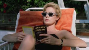 ‘Strangers Kiss’ Blu-Ray Review – A Palpable Look At The Murky Lines Of Professional Intimacy A person wearing sunglasses and a black top reclines on an outdoor lounge chair, reading a book titled "And Now Good-Bye.