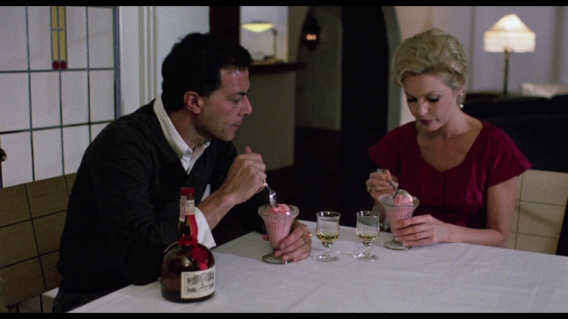 Two people sit at a table eating from dessert dishes. A bottle and three glasses are also on the table.