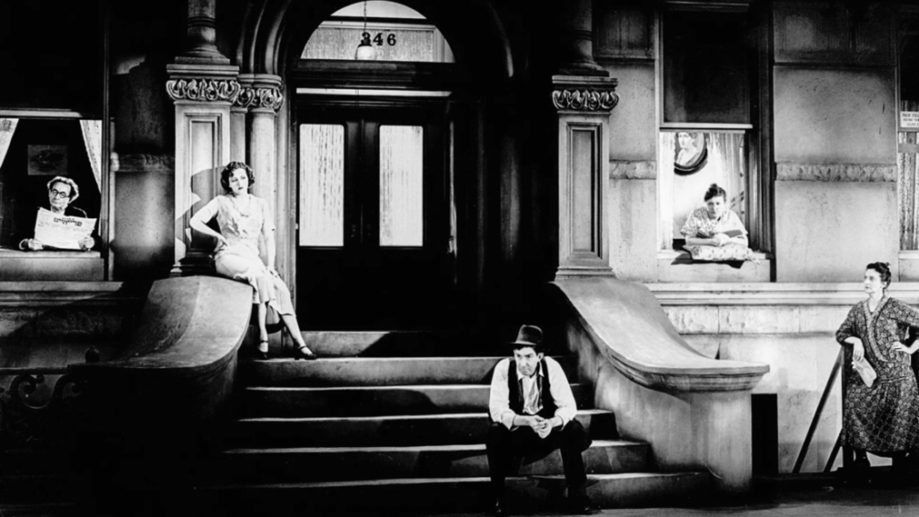 Black-and-white photo of four people outside a building with arched doorways and steps. One person is seated on the steps, two women lean out of windows, and another woman stands with a broom.