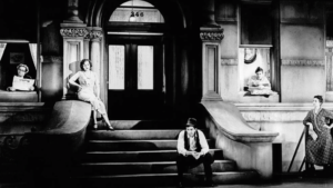 ‘Street Scene’ (1931) Blu-Ray Review – Sweltering Pre-Code Drama Boils Over Black-and-white photo of four people outside a building with arched doorways and steps. One person is seated on the steps, two women lean out of windows, and another woman stands with a broom.