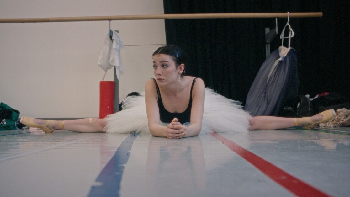 A ballet dancer in a white tutu stretches on the floor with legs in a wide split, leaning forward on her elbows, in a dance studio with costumes hanging in the background.
