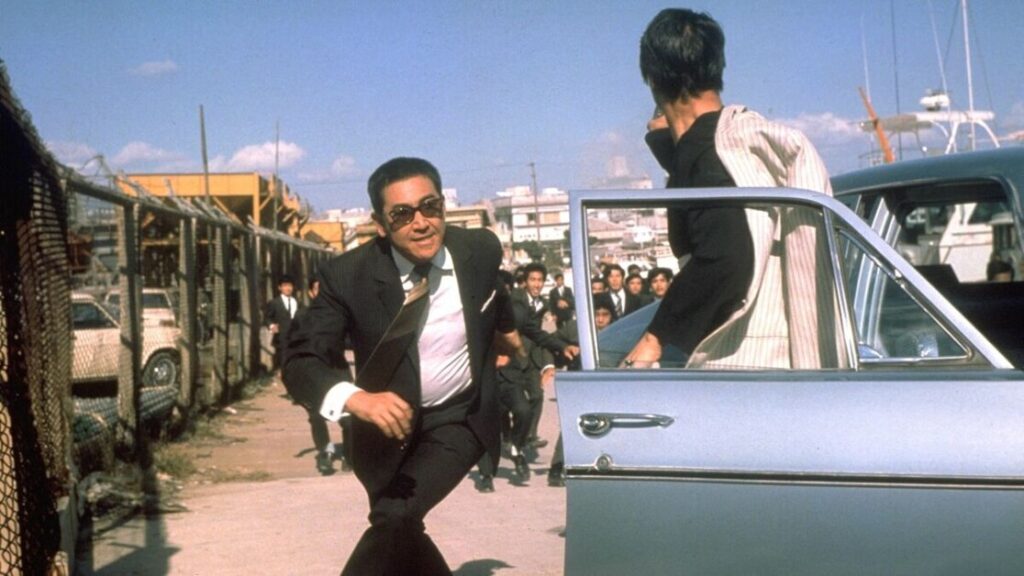 A man in a suit runs towards an open car door while another man stands near the door, set in a busy area with a crowd in the background.