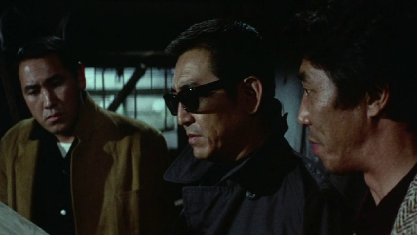 Three men in a dimly lit room have a serious discussion. The middle man wears sunglasses and a dark coat, and the others listen attentively.