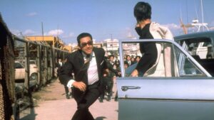 A man in a suit runs towards an open car door while another man stands near the door, set in a busy area with a crowd in the background.