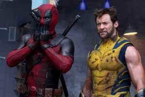 Two costumed characters stand side by side; one in a red and black suit with hands on hat face, the other in a yellow suit looking forward.