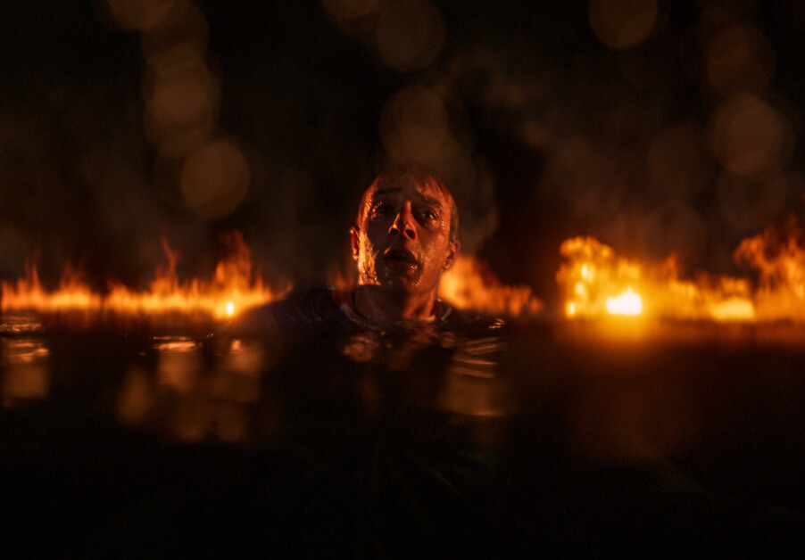 Person partially submerged in water, surrounded by flames, with a concerned expression in a dark setting.