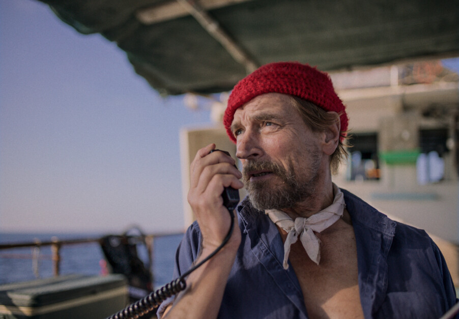 A man wearing a red beanie and blue shirt speaks into a handheld radio on a boat. The background shows part of the boat structure and the ocean.