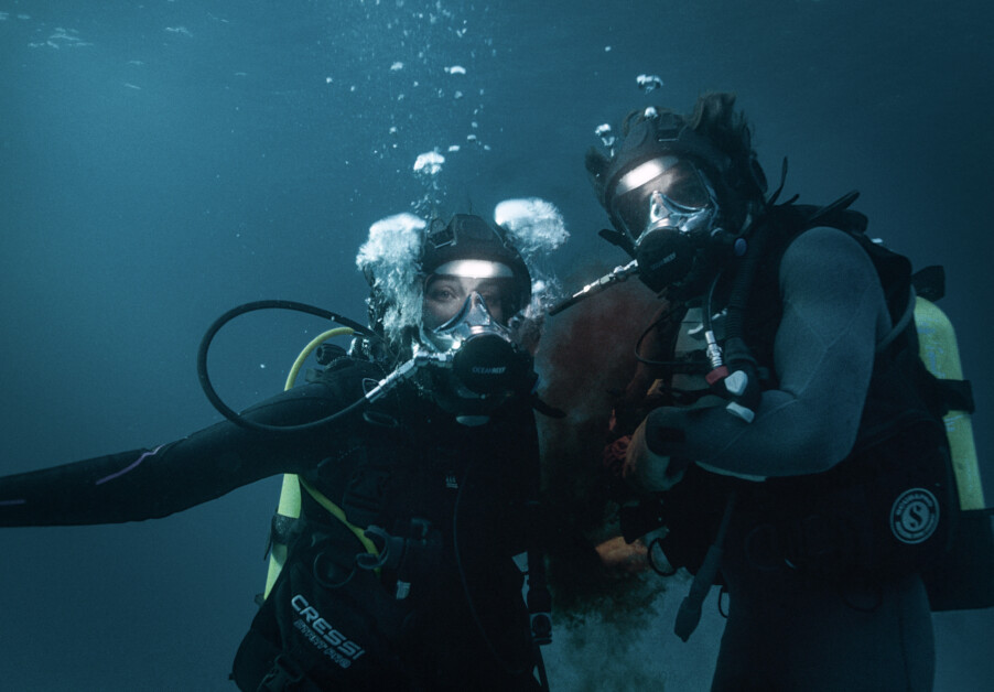 Two scuba divers underwater wearing full scuba gear and masks with bubbles rising around them.