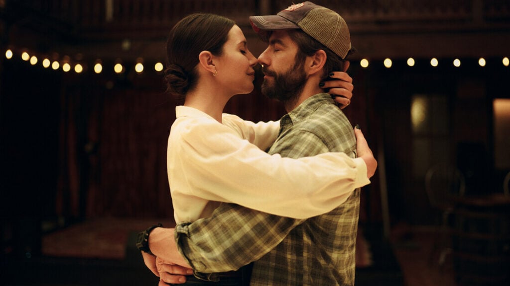 A couple embraces closely with their arms wrapped around each other in a warmly lit room. The woman wears a white top and the man is in a plaid shirt and cap. String lights are visible in the background.
