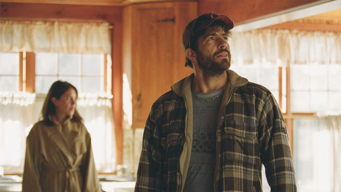 A man in a flannel shirt and cap looks upward inside a wooden room, while a woman in a coat stands in the background.