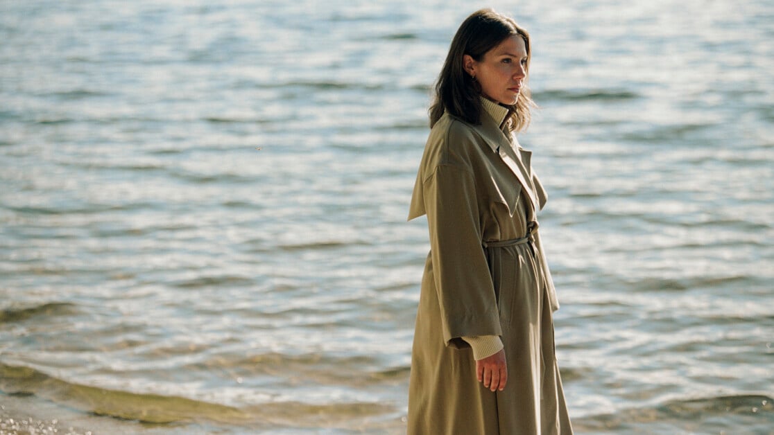 A person wearing a long coat walks along the shore of a body of water, facing to the right.