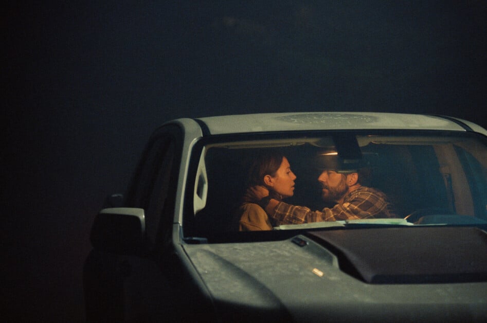 A man and a woman sit in a dimly lit car, facing each other closely. The woman's hand is on the man's neck as they share an intimate moment.