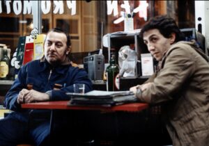 Two men sitting at a red table, one in a blue outfit and the other in a brown jacket, with various items including drinks, a radio, and papers on the table, in what appears to be a bar or cafe.
