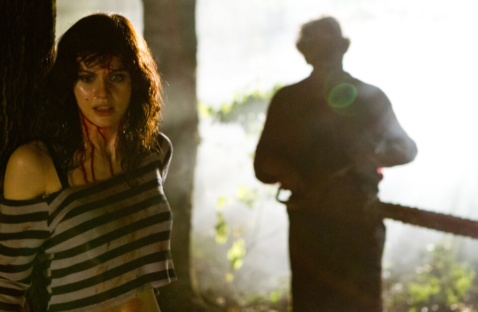 A frightened woman with blood on her face and wearing a striped shirt stands in the foreground. Behind her, a silhouette of a figure holding a chainsaw appears in the misty background.