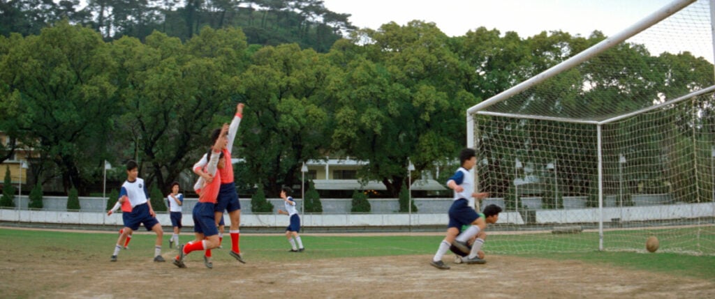 A group of youths plays soccer on a field. One player in a red vest jumps near the goalpost while another is on the ground. The ball is seen moving toward the net. Trees and buildings are in the background.