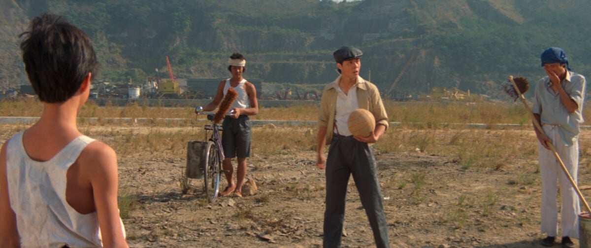 Four men stand outdoors on a dirt area with a hilly backdrop; one holds a basketball, another is by a bicycle, and one smokes a pipe.