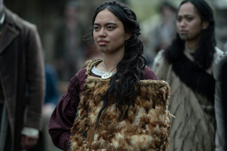A woman with traditional facial markings and wearing a feathered garment stands outdoors, with another similarly dressed person in the background.