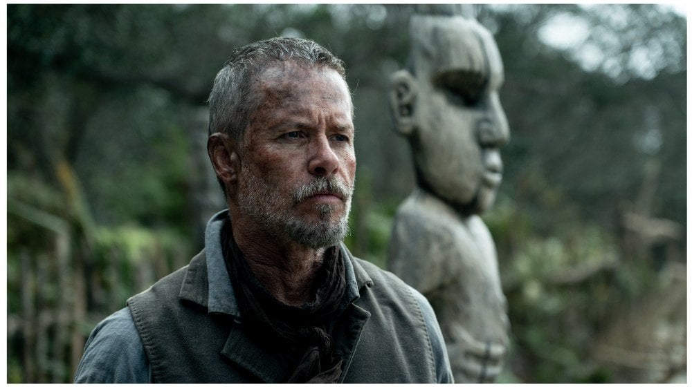 A man with a short gray beard and messy hair stands outdoors in front of a large, carved wooden statue with a forested background.
