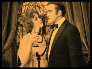 A black-and-white image of a formally dressed man and woman from an old film. She holds a glass and looks at him, while he gazes back intently. The background has draped curtains and foliage.