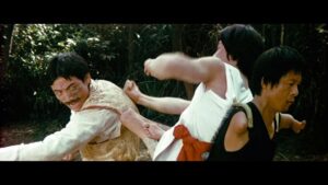 Three individuals engaged in martial arts combat outdoors, with one person in white delivering a punch while another in black dodges, and the third in white looks back. Trees and foliage in the background.