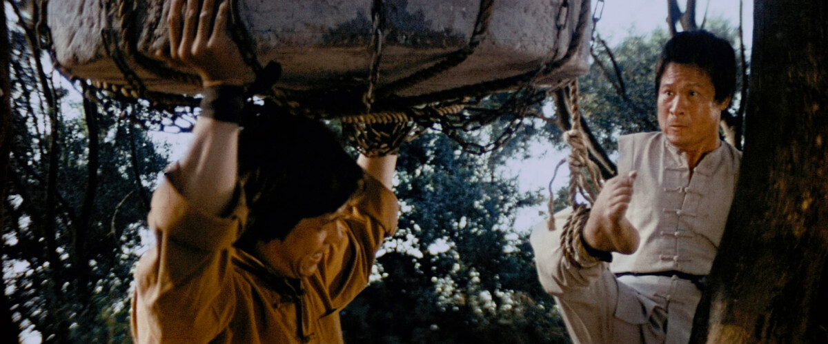 Two people in traditional attire, with one holding up a large stone object using a rope net, while the other watches from nearby. Trees and foliage are visible in the background.