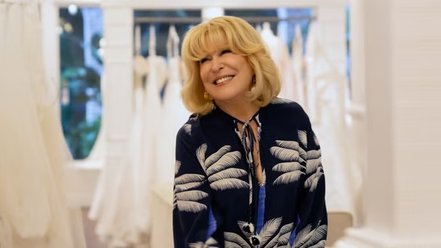 A smiling woman with blonde hair stands indoors, wearing a dark blouse with a white leaf pattern. White dresses on hangers are visible in the background.