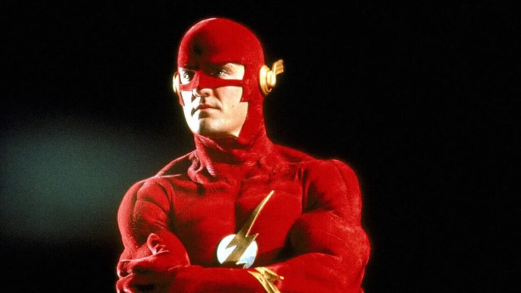 A person in a red superhero costume with a lightning bolt symbol on the chest and gold accents on the mask stands with arms crossed against a dark background.