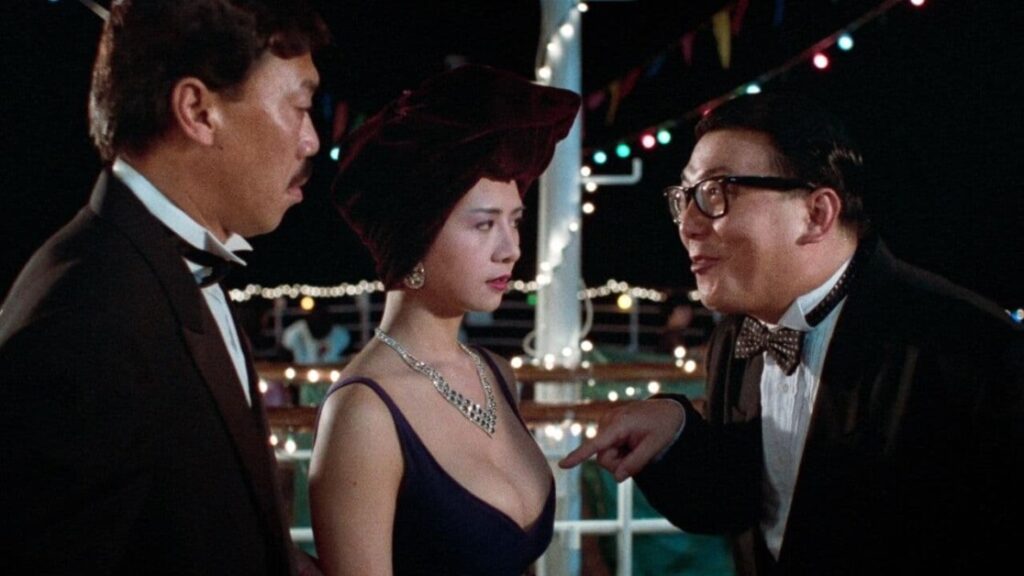 Three people dressed formally engage in conversation at a nighttime event decorated with string lights and colorful triangular flags.