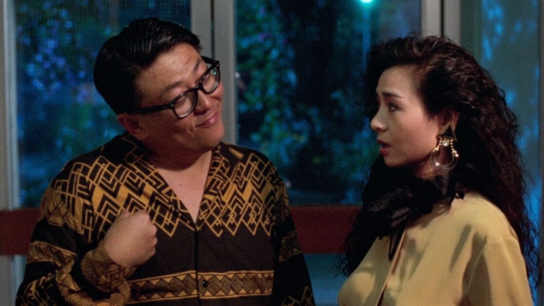 A man wearing glasses and a patterned shirt gestures while talking to a woman with long curly hair and statement earrings, who appears to be listening attentively.