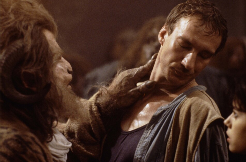 A creature with horns grasps a man's neck, while another person looks on in the background.