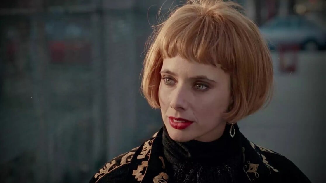 A person with short, auburn hair and red lipstick is looking to the side, wearing a black outfit with a patterned jacket.