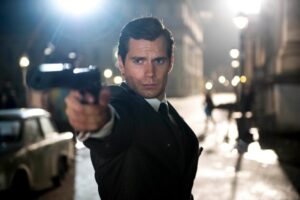 A man in a suit aims a handgun directly forward on a dimly lit street at night. Vintage car and blurred figures are visible in the background.