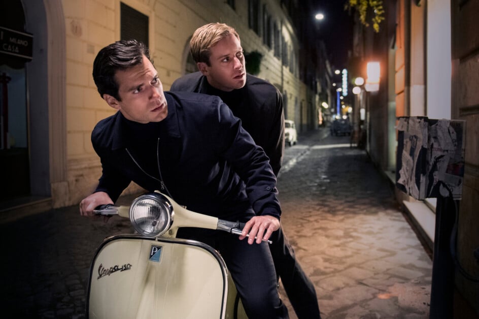 Two men in black clothing ride a cream-colored Vespa scooter down a dimly lit cobblestone street at night, appearing to look back over their shoulders with concerned expressions.