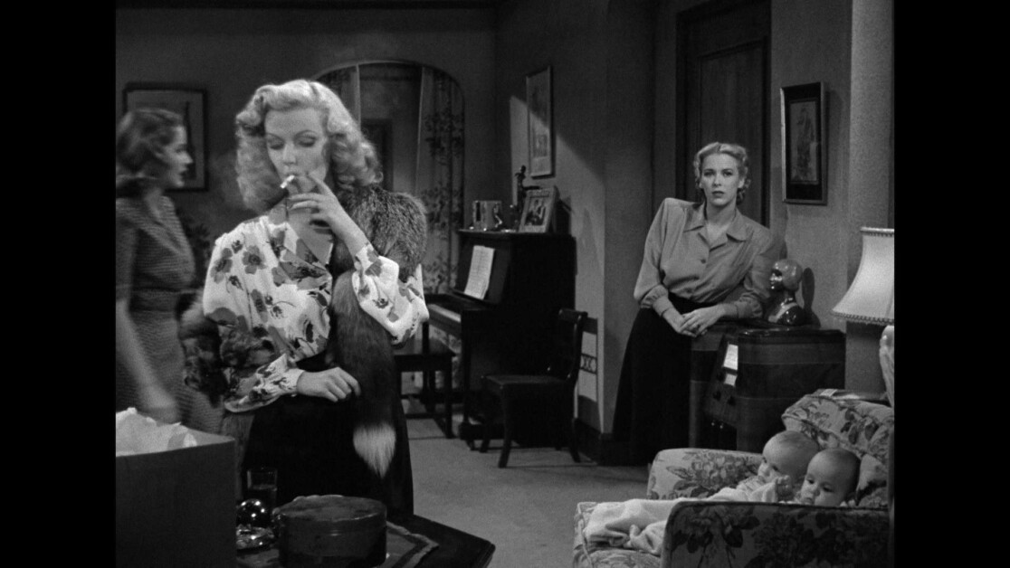 Three women are in a living room. One smokes a cigarette, another stands by a doorway, and the third is blurred in motion. Two babies sit in a bassinet on the right side of the image.