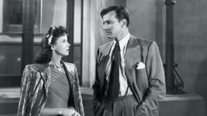 A man and a woman stand facing each other in formal attire. The image is in black and white and has a classic, vintage look.