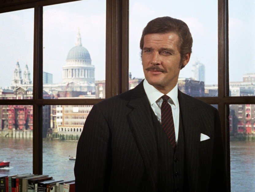 A man in a suit and tie stands by a large window, with a view of a cityscape including a dome and river in the background.