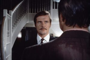 A man with a mustache, wearing a suit and tie, stands in front of a staircase, facing another person whose back is to the camera.