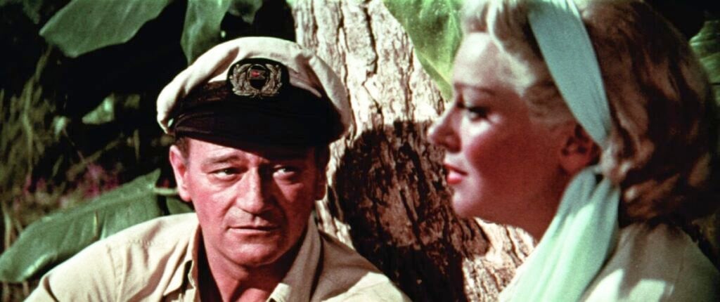 A man in a captain's hat and a woman with blonde hair and a blue headscarf sit beside a tree.