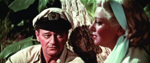 ‘The Sea Chase’ Blu-Ray Review – John Wayne Breaks Out Of His Comfort Zone In Seafaring War Tale A man in a captain's hat and a woman with blonde hair and a blue headscarf sit beside a tree.