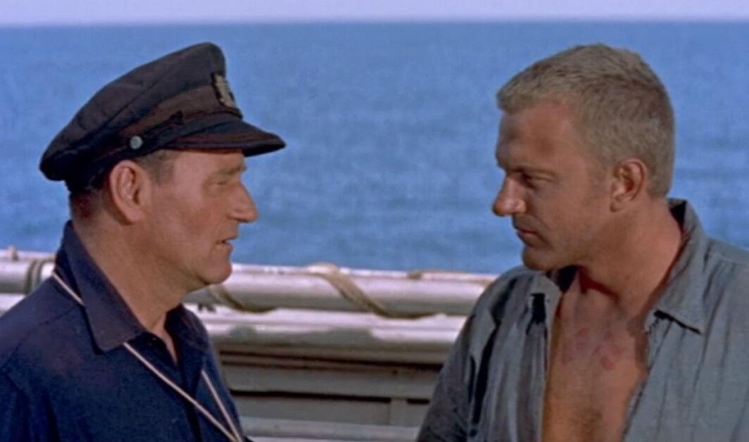 Two men stand on a boat, engaged in conversation. The man on the left wears a dark uniform and cap, while the man on the right, shirt partially unbuttoned, faces him. The ocean is visible in the background.