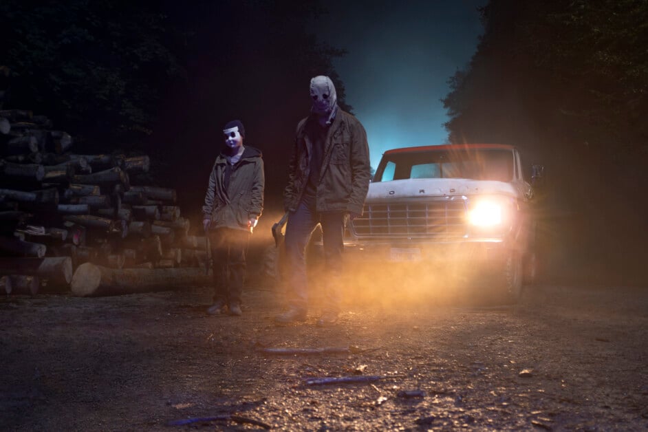 Two people wearing masks stand on a dirt road at night in front of a truck with its headlights on, surrounded by trees and logs. Fog and eerie lighting enhance the suspenseful atmosphere.